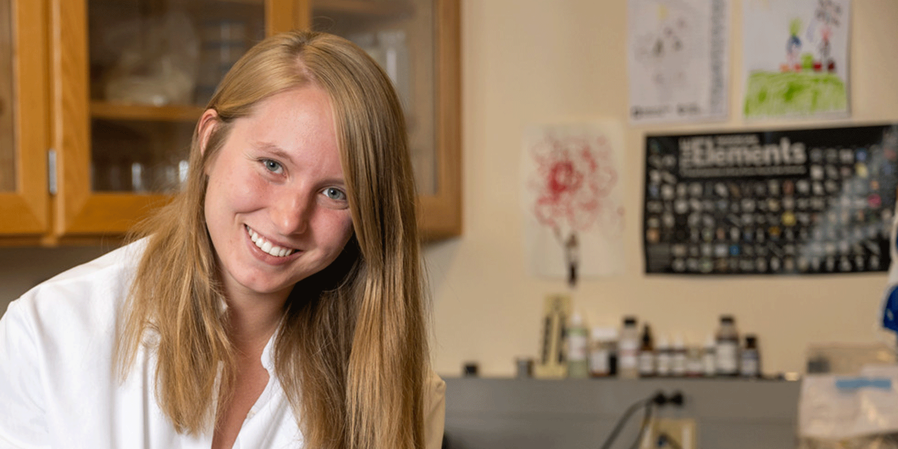 A white female student with long blond hair poses in a laboratory space. She wears a white lab coat. Behind her are brown cabinet doors, medical bottles, and various posters on the wall, including the Periodic Table of Elements.