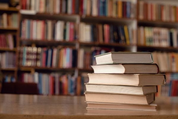 A stack of six books sits on a table in the library. The spines are facing away and cannot be read. In the background, shelves of colorful books are visible.