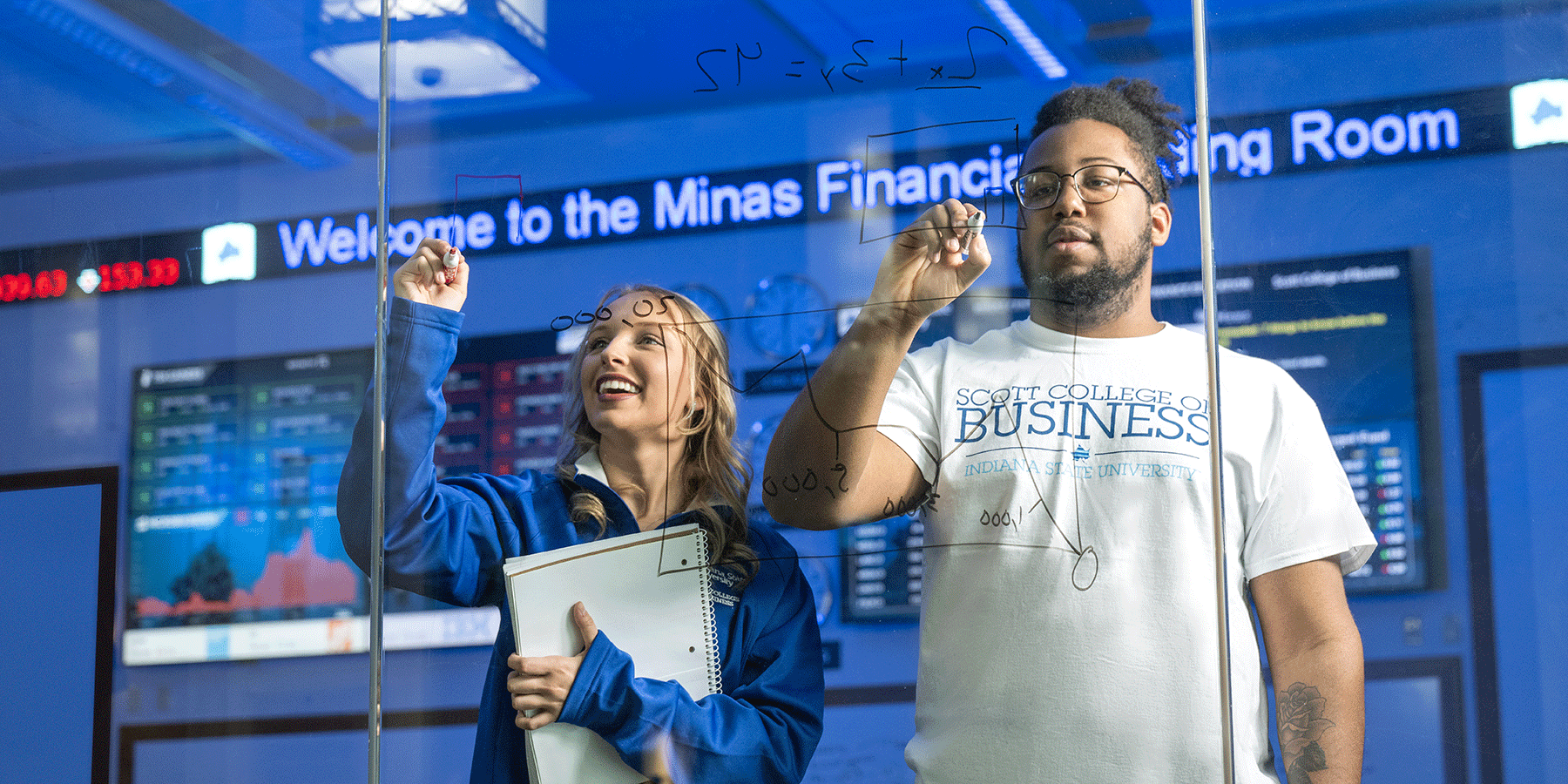 Two students write on a clear dry erase board with news ticker in background