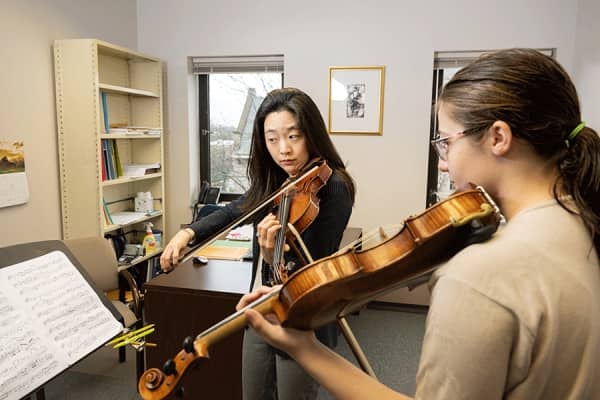 An Asian female professor with black shirt plays a violin while looking at sheet music as a White female student follows along.
