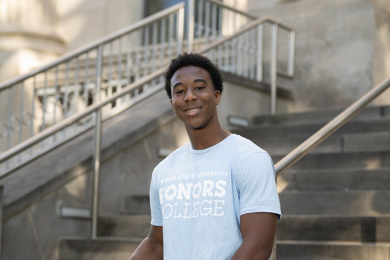 A black male student poses outside on grey steps and a silver railing. He wears a light grey T-shirt with Indiana State University Honors College in white lettering. 