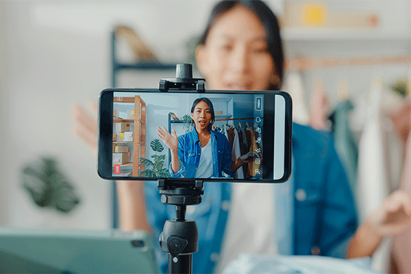 Young woman in a blue jacket recording a video on a smartphone mounted on a tripod, waving while speaking to the camera in a home studio setting.