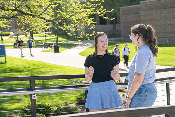 Two college students, both women with dark brown hair, talk and smile on a sunny Indiana State University walkway, with other students walking in the background and green trees, grass, and brick buildings surrounding the scene.