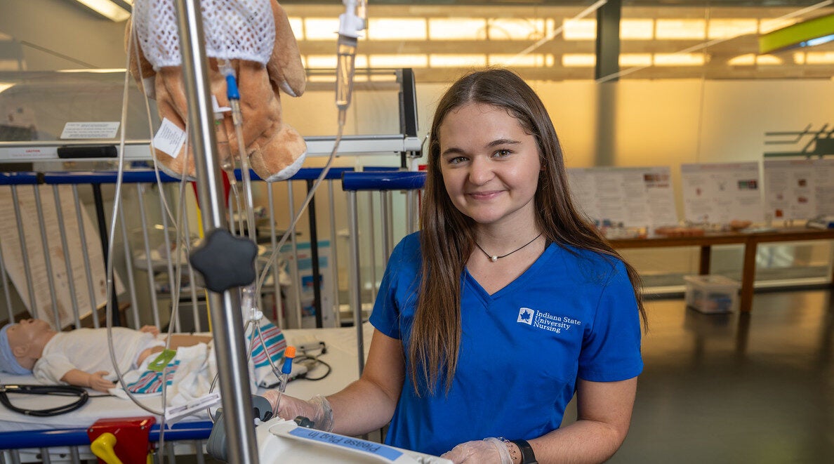 A white female with long brown hair in blue nursing scrubs labeled "Indiana State University Nursing" stands next to medical equipment. In the background, a medical mannequin lies on a bed, surrounded by various medical instruments and devices.