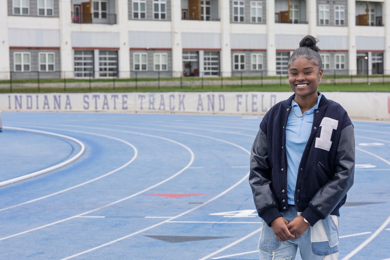 A Black female stands on a blue track field in front of a building with multiple windows. She is wearing a dark blue varsity jacket with an "I" on the right side and light blue jeans. In the background, the words "Indiana State Track and Field" are written on a wall.