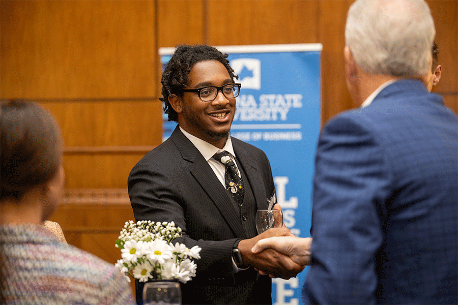 A Black male student in a black suit is shaking hands with another person in a blue suit. They are standing in front of a blue banner that reads Indiana State University. There are white flowers on the table in the foreground, and two other people are partially visible on either side of the image.