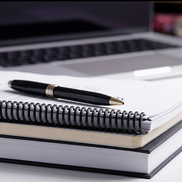 A close-up view of a workspace featuring a laptop in the background, an open spiral-bound notebook in the foreground, and a pen resting on the notebook. 