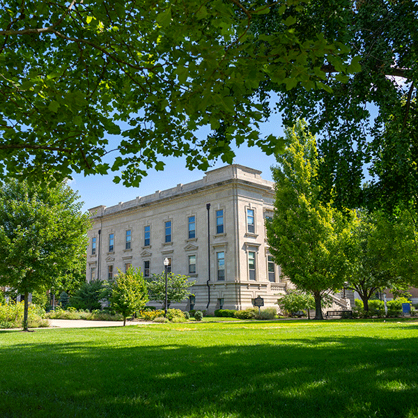 A large, classical-style building with multiple windows, surrounded by green trees and a lawn. The sky is clear and blue. 