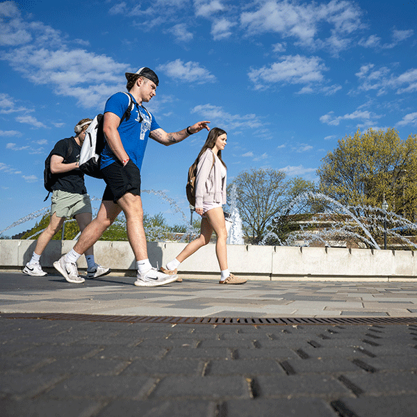 Three people walk along a paved pathway with a fountain in the background. The sky is mostly clear with a few scattered clouds, and trees are visible in the distance. 