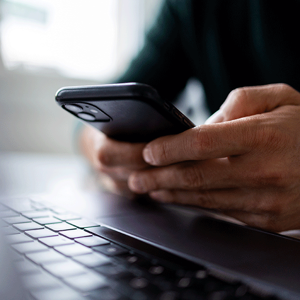 A close-up photo of a person’s hands holding and using a smartphone, with part of an open laptop visible in the foreground.
