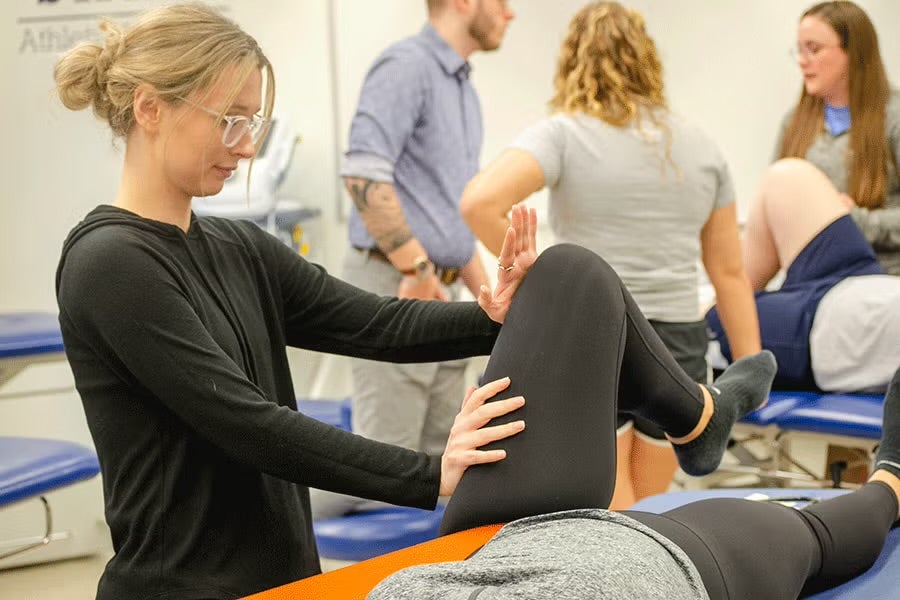 A white female with blonde hair pulled back into a bun is assisting a patient with a leg exercise on an examination table. The woman is holding the patient's bent knee and ankle, guiding the movement. In the background, other people are working with patients on similar tables. The room appears to be a clinical setting with medical equipment and blue padded tables. 