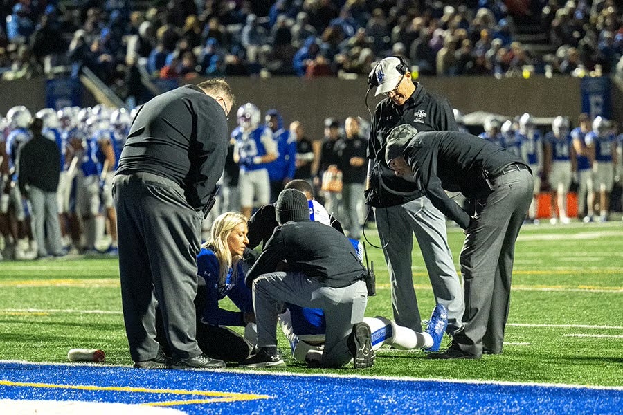 A group of five people, including medical staff and coaches, are attending to an injured football player on the field. The player is lying on the ground near the sideline with one leg extended. The background shows other players and spectators in a stadium setting.