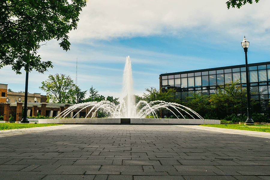 A large fountain with multiple streams of water shooting upward and outward is set in a landscaped area. It is surrounded by a paved walkway, trees, and buildings, including a modern glass building on the right and a brick building on the left. The sky is mostly clear with some clouds, and lampposts are visible near the fountain.