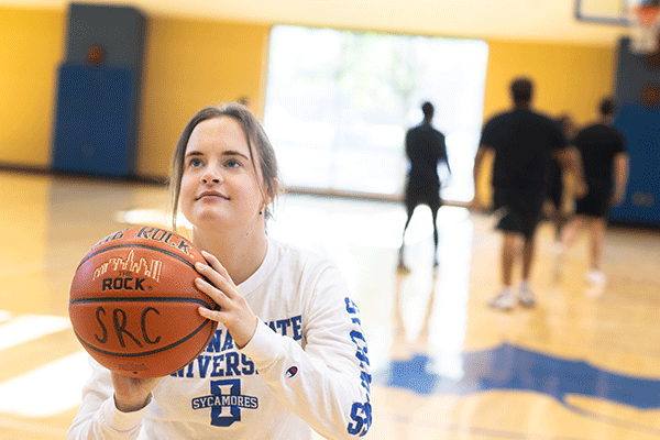 A young woman stands in a gym, confidently holding a basketball with a focused expression. 