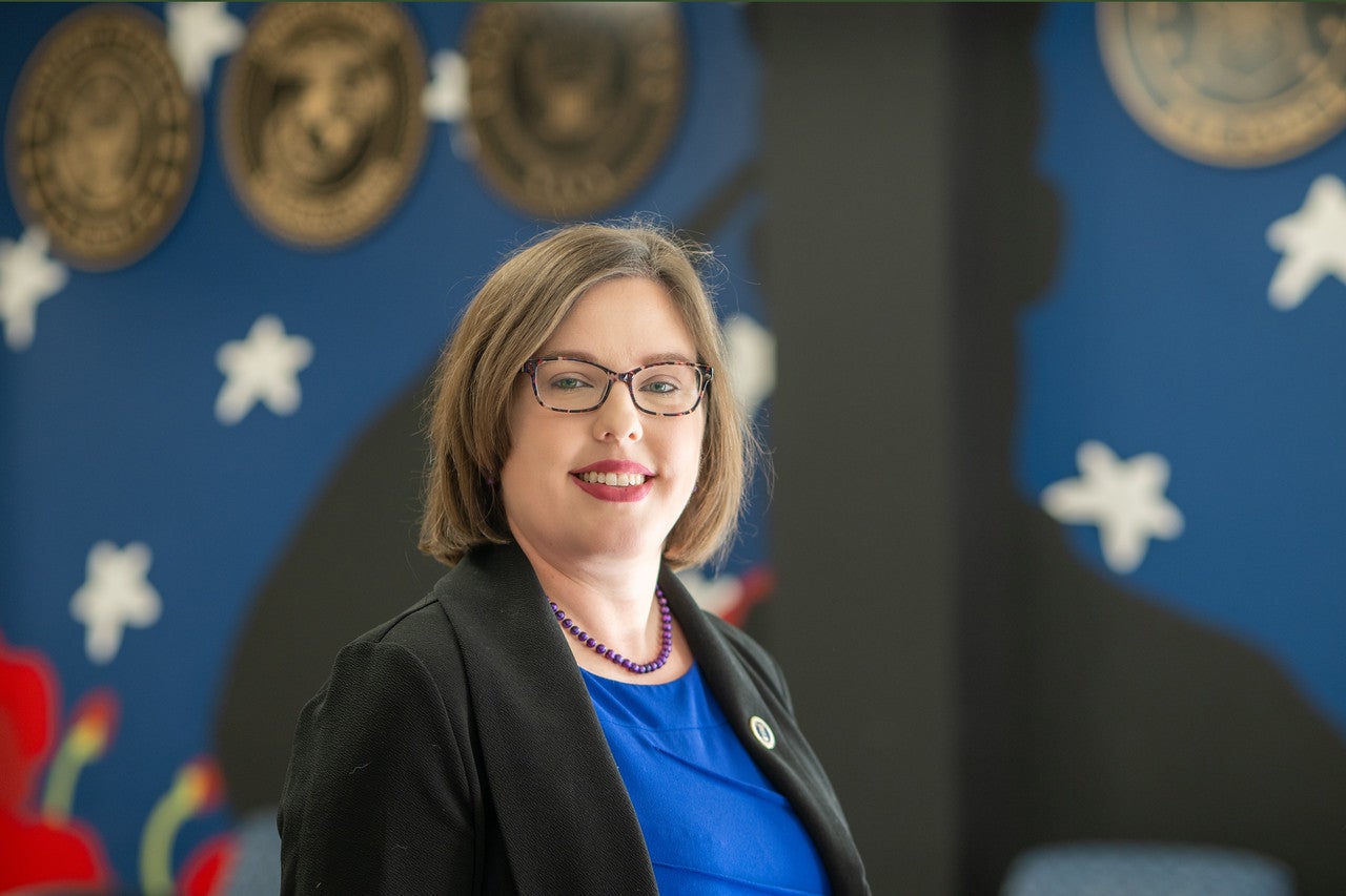 Woman with shoulder-length brown hair stands in front of a patriotic mural featuring stars and military service emblems, including Army, Marine Corps, Navy, Air Force, and Coast Guard seals. She wears a blue shirt with a black blazer jacket. 