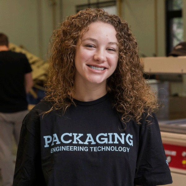 A smiling young woman with curly, shoulder-length brown hair stands in a workshop. She wears a black t-shirt that reads "Packaging Engineering Technology." 
