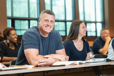 A middle-aged white male with short greying hair and a grey t-shirt sitting at a table in a classroom looks toward the camera and smiles with a diverse group of classmates in the background.  