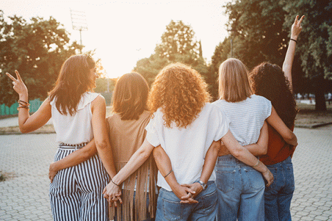 A group of five women are seen standing from the back. They are holding hands and have their arms around each other as they look toward a sunset framed by green trees in the background. The women wear an assortment of jeans, t-shirts, and other summer clothing, and their hair has various styles and colors. 