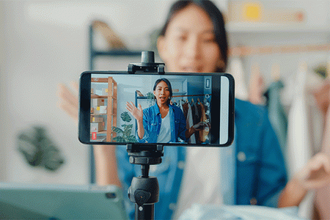 Young woman in a blue jacket recording a video on a smartphone mounted on a tripod, waving while speaking to the camera in a home studio setting.