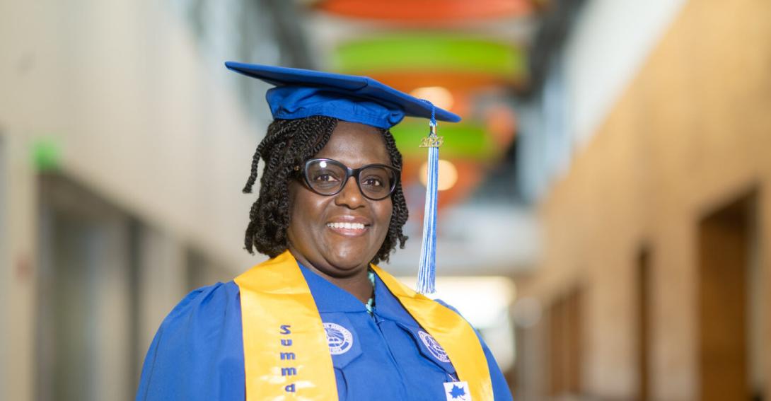 A Black female with black glasses wearing a blue graduation cap and gown with a yellow stole stands in an indoor hallway with colorful ceiling panels. 