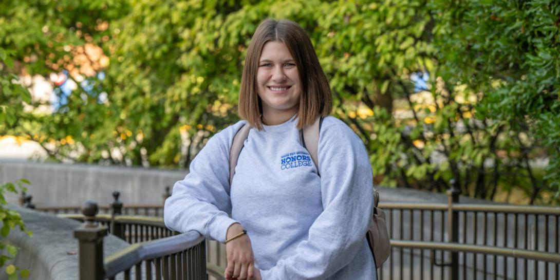 A white female student with shoulder-length brown hair stands outdoors near a metal railing, wearing a light grey sweatshirt with the text “HONORS COLLEGE” and carrying a backpack. The background features green trees and partially visible buildings through the foliage. 
