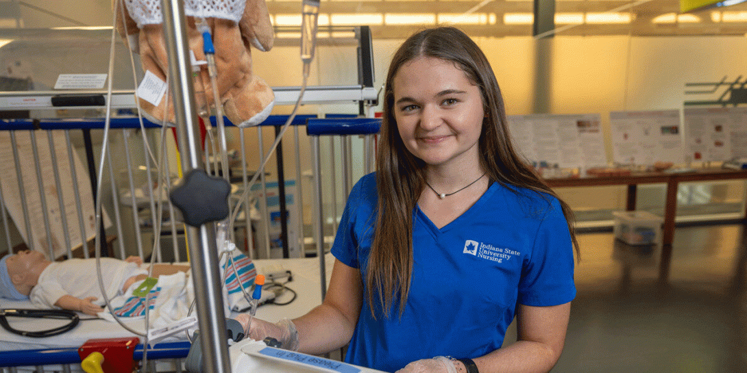  A white female student with long brown hair wears blue scrubs and stands next to a hospital crib with medical equipment. The crib contains a doll, and various medical instruments are attached to it. In the background, there are informational posters and a table with additional medical supplies.
