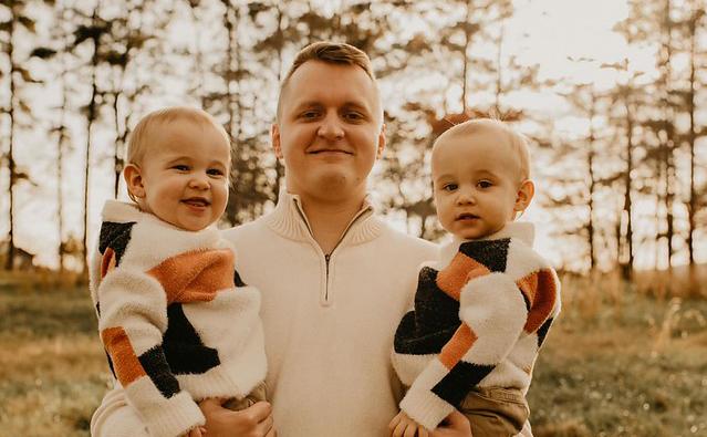 In the center, a white male with short brown hair holds two children, one on each side. The background features trees and a grassy area. The children wear matching sweaters with geometric patterns in black, white, orange, and beige colors. The adult wears a white zip-up sweater. 