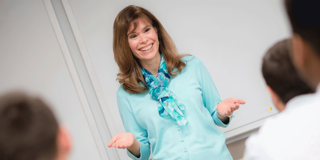 A white female with long brown hair wears a light blue dress shirt and a patterned scarf. She stands in front of a whiteboard in a classroom setting. Three individuals are visible from behind in the foreground, appearing to listen.